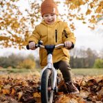 Smiling child in yellow jacket rides a bike through fallen autumn leaves in a park.